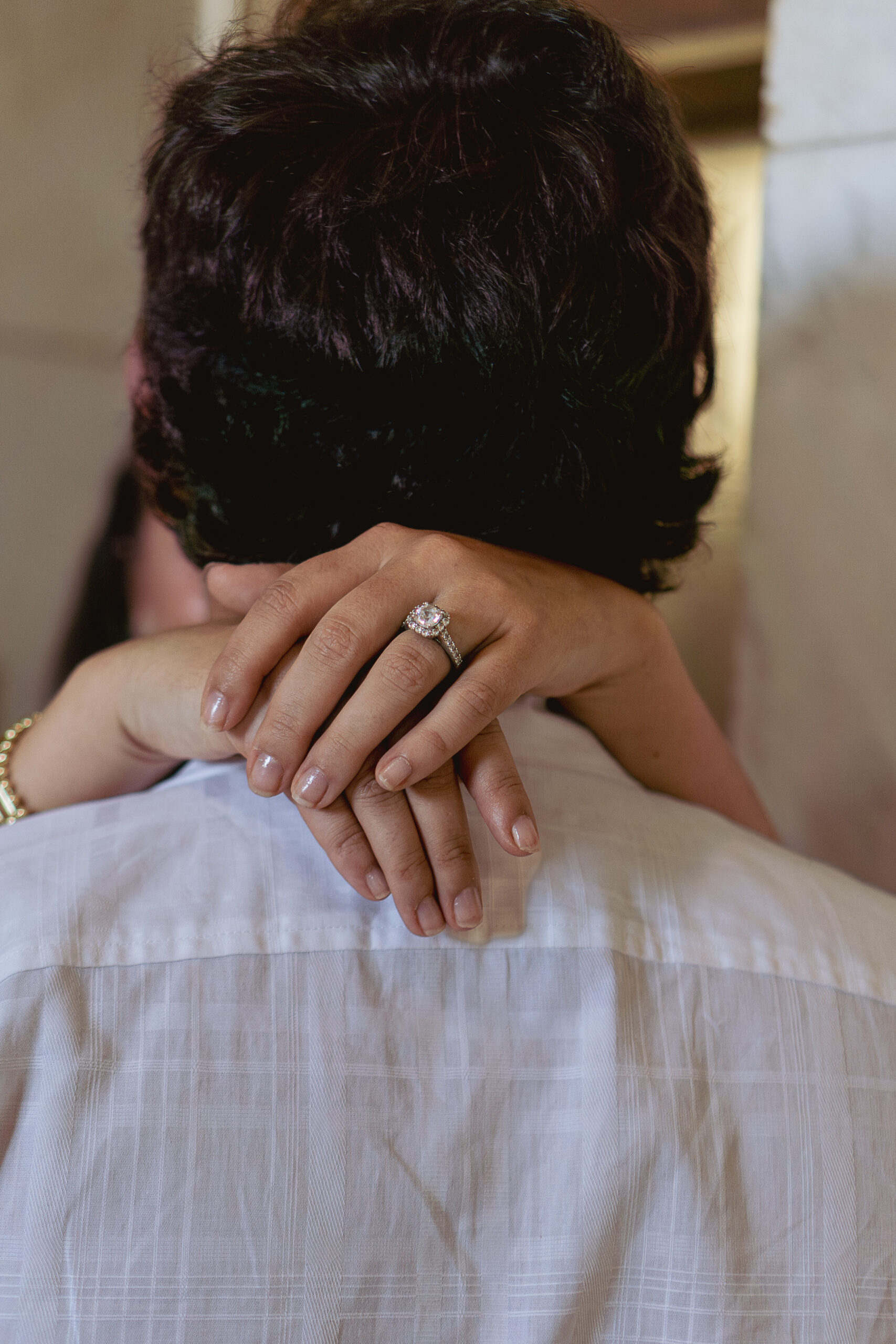 Couple facing each other, man's back to camera. Woman's arms around neck, focus on engagement ring.